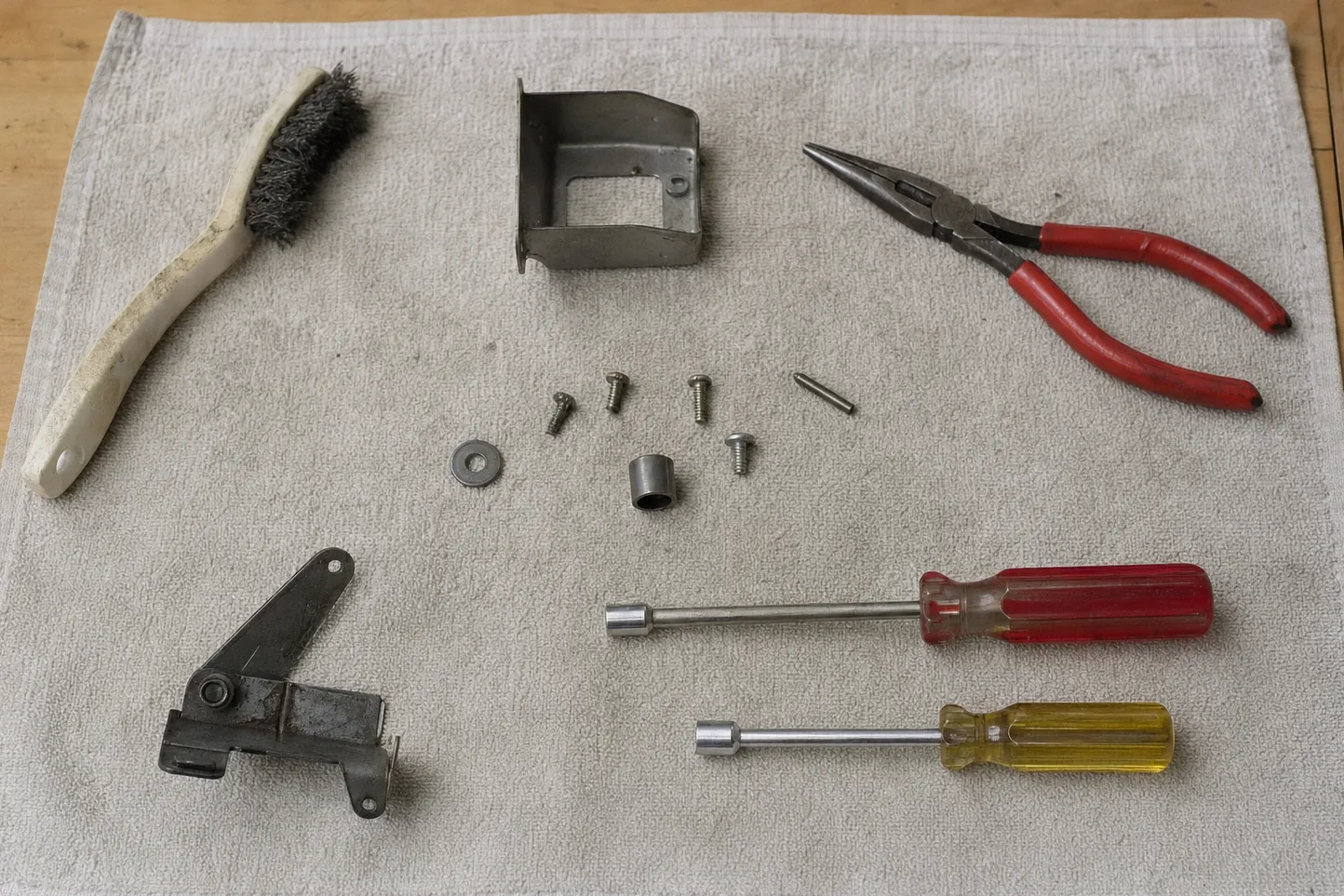 Wire brush, needle-nose pliers, two nut drivers, a door hinge assembly and loose fasteners on a work cloth
